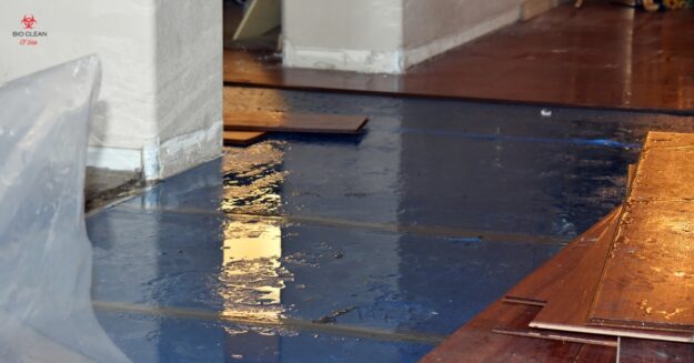 A blue floor is partly covered in water from a leak. Plywood sheets are stacked to the side, and a plastic barrier is visible on the left—ideal for flood restoration by a professional restoration service.