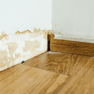 Damaged wall with peeling paint at the base, next to a wooden skirting board and wood-patterned floor—ideal for professional water damage restoration Utah services.