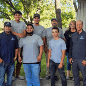 A group of eight people, six men and two women, stand outdoors in front of trees and a building, smiling at the camera in matching gray Fire & Smoke Damage Cleanup shirts.